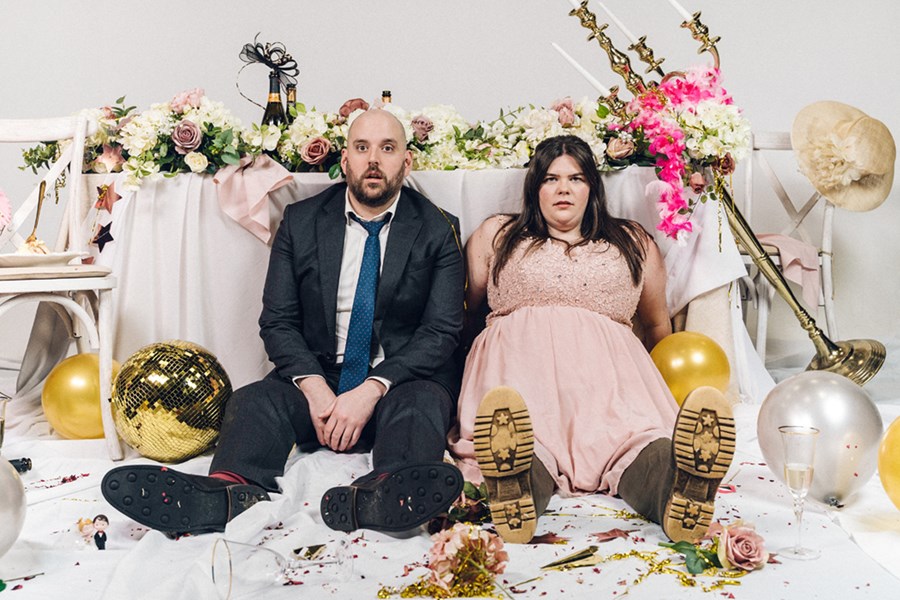A man and woman sit on the floor with dazed expressions. The man is wearing a suit with a messy tie and the woman is wearing a soft pink floor-length dress and wellies. There is a messy table behind them covered in empty bottles, ruined flower displays and a large candelabra that has fallen over. On the floor around them are balloons, confetti and a broken disco ball.