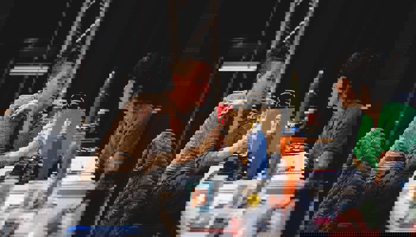 Two women sit at opposite sides of a table covered in water bottles and coffee cups. One woman has her mouth slightly open and is pointing to something on the table. She looks directly at where she is pointing. The other woman is looking directly towards her.