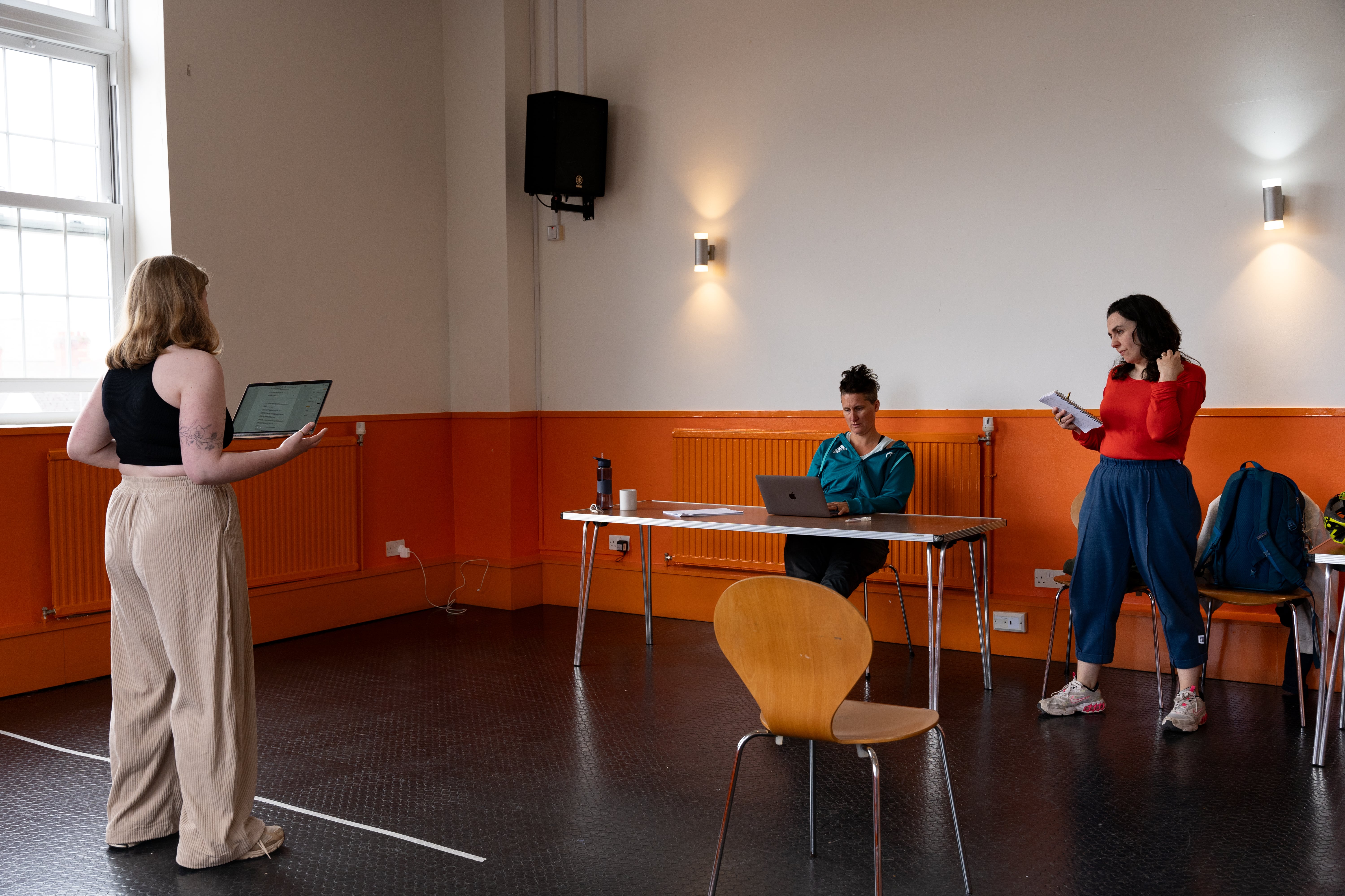Image from Bren Calon Fi rehearsals with Lowri Morgan, Rhiannon Mair, and Bethan Marlow. The rehearsal space has white walls with a painted orange border around the bottom. Bethan is sat at a table with her laptop looking at Lowri who has her back to the camera. Rhiannon stands next to Bethan.