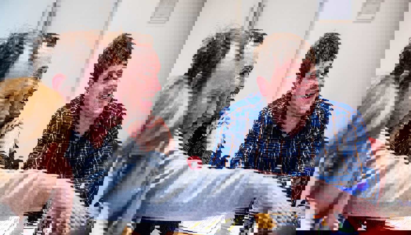 4 people sat a table. We see 3 men sitting next to each to each other, and the back of one woman's head who is facing away from the camera. The men are dressed informally and are all looking at the same point off camera. There is paper on the table in front of them.