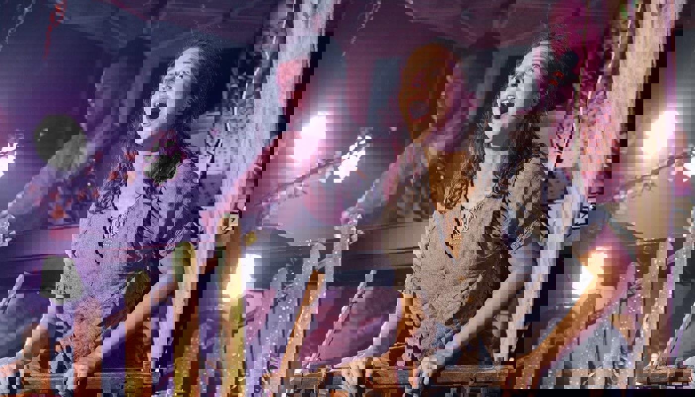 A woman dressed in grey sits in a small wooden cart. She is singing. Two musicians are in the background - a man with a guitar and a woman singing and playing keyboard.