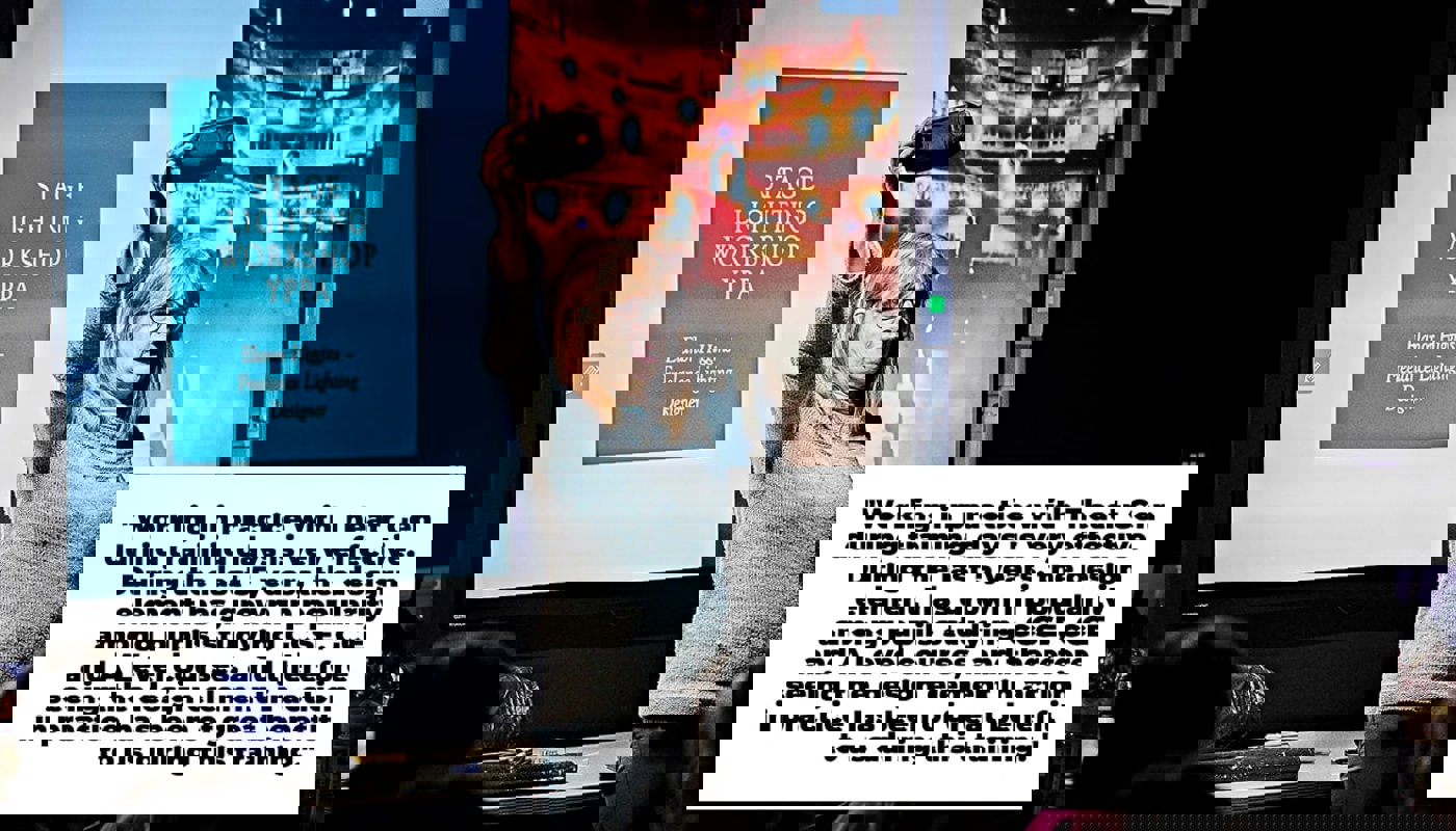 Woman stands in a darkened room in front of a projector screen. Presentation slide on the screen behind her reads 'Stage Lighting Workshop'. She is holding a phone above her face and the torch is on, illuminating her face. She is looking out in front of her, not into the camera. People are sat around her on chairs.