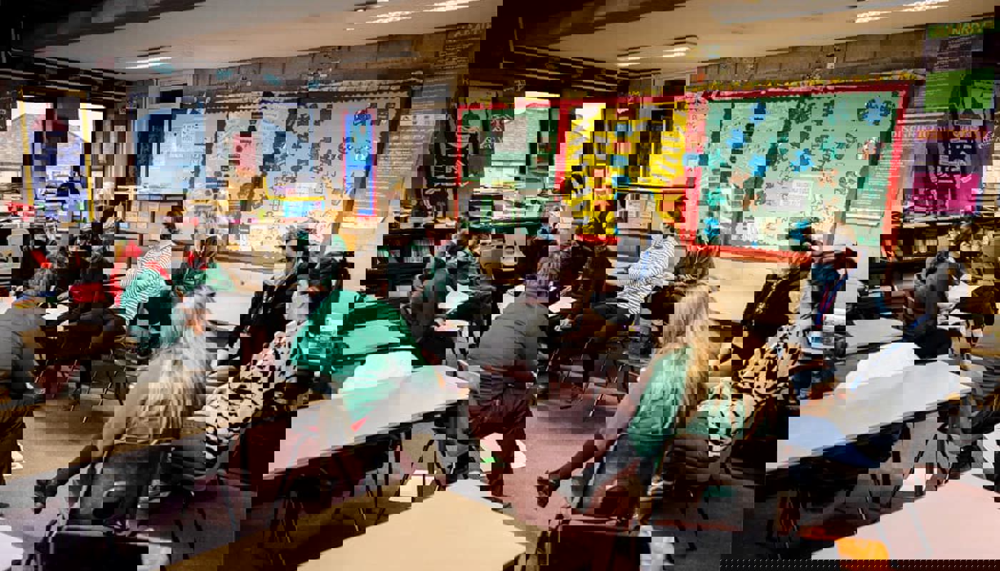 Interior of a busy classroom. The walls are covered in multicoloured informational posters and pupils’ work. A mix of pupils and teachers are sitting in a circle together. Everyone is looking at one of the individuals who is mid conversation. Many of them are wearing face masks.