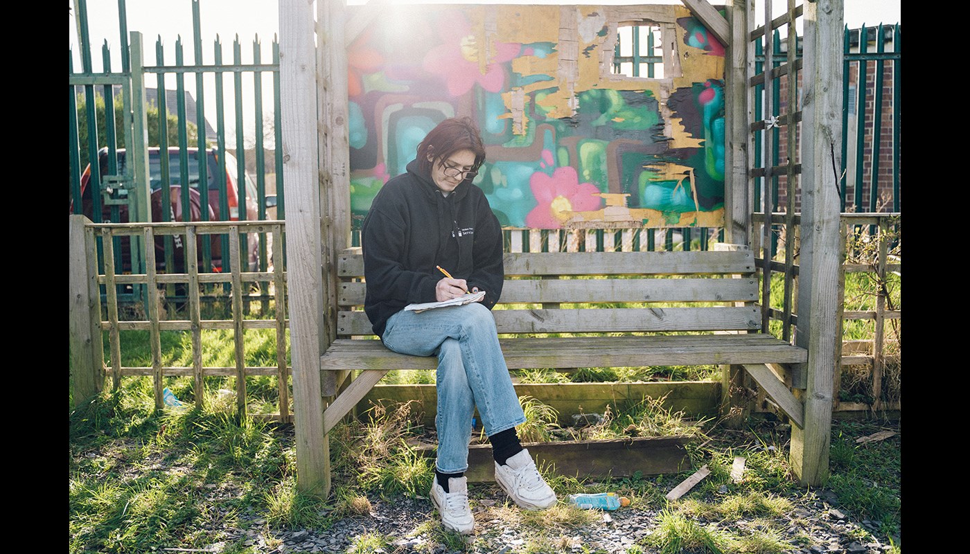 Woman sits outside on a wooden bench. The sun is shining and she is holding a pen in one hand while resting a notebook on her legs. She holds the pen to the paper and looks down.