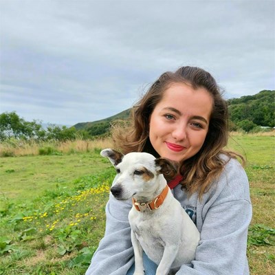A photo of Sioned Erin. She is a young white woman with shoulder length brown hair. She is smiling and looking directly into the camera. She is sat outside near some hills on a patch of grass holding a small terrier dog.