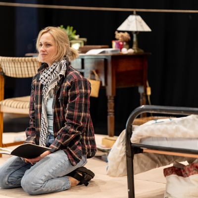 Rehearsal photo for Fy Enw i yw Rachel Corrie. A woman wearing a keffiyeh, check shirt and jeans is kneeling on the floor and holding a notebook in her hands.