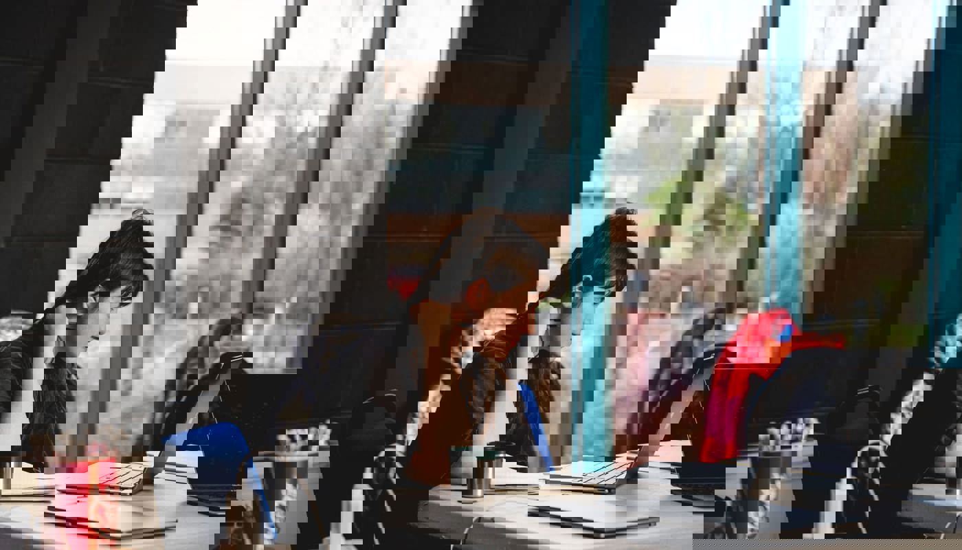 Young woman sat at a table. She is looking down at a laptop in front of her. She has a pen in her hand and there is an open notebook next to the laptop.