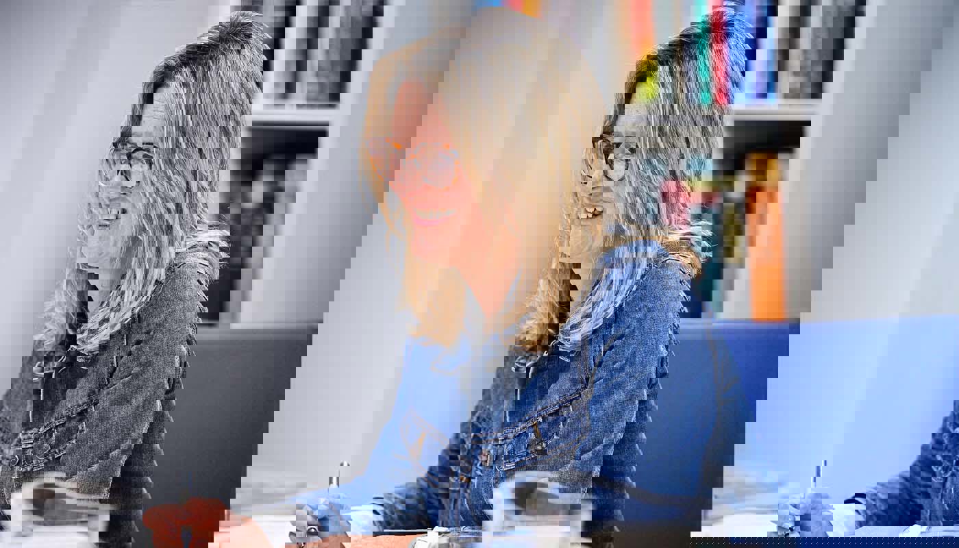 6.	Middle-aged woman sits at a table. Her arms are resting on the table and she has a pen in one hand. She is smiling with her mouth open and looking off camera.