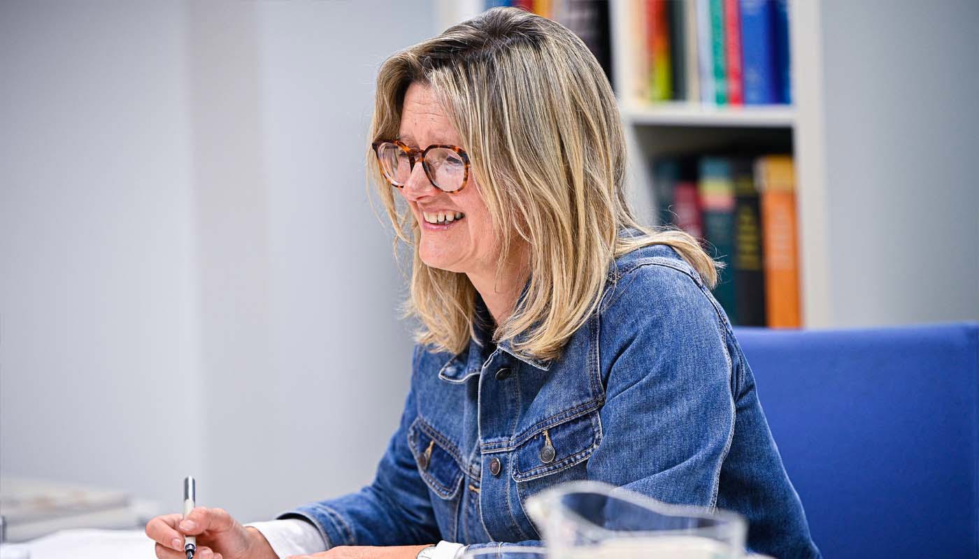 6.	Middle-aged woman sits at a table. Her arms are resting on the table and she has a pen in one hand. She is smiling with her mouth open and looking off camera.